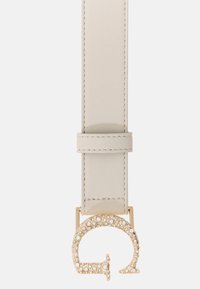 Hellbeiger Ledergürtel mit strukturierter Oberfläche, versehen mit einer runden goldfarbenen Schnalle, die mit Strasssteinen und einem stilisierten Logo verziert ist.