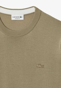 Oliivinvihreä neulepaita, jossa on pyöreä kaula-aukko ja ribbineulereunat. Rinnassa on brodeerattu Lacoste-logo. Vakiokokoinen malli.