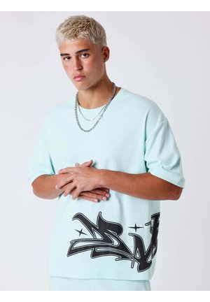 Giovane uomo con capelli corti decolorati, indossa una camicia azzurro chiaro con stampa nera graffiti e collane di catena argentate, mani giunte.