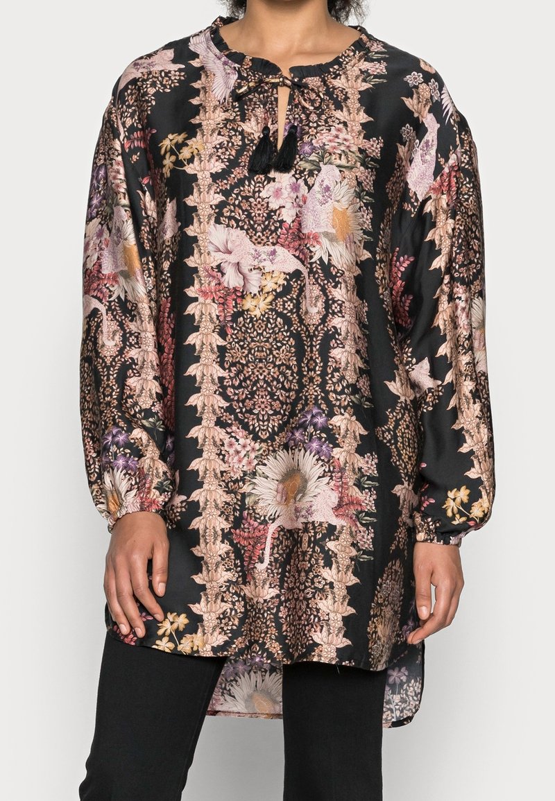 Femme portant une tunique noire à manches longues avec des motifs floraux et d'oiseaux roses, beiges et violets et un col à nouer avec des pompons noirs.