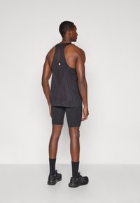 Μαύρο αθλητικό φανελάκι με σχέδιο racerback, υφή υφάσματος και μια μικρή λεπτομέρεια λογότυπου. Συνδυάζεται με μαύρες συμπιεστές σορτς και παπούτσια.