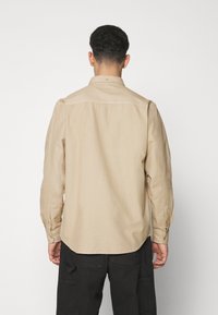 Camisa de manga larga beige con cuello button-down, textura suave y dobladillo curvado, combinada con pantalones cargo negros.
