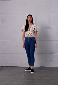 Blusa floral branca com mangas esvoaçantes, combinada com jeans azuis de cintura alta e sapatilhas pretas. O fundo é uma parede cinza lisa.