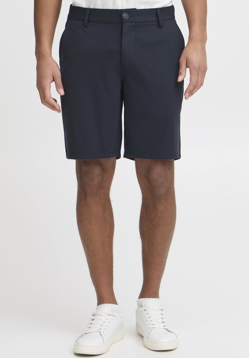 Marineblå shorts laget av et glatt stoff, med knappelukking, frontlommer og en skreddersydd passform. Båret med hvite joggesko.