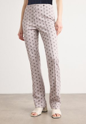Femme portant un pantalon slim à motifs taille haute et des chaussures blanches à talons blocs ouverts, debout sur un sol gris.