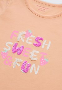 Chemise couleur pêche avec le texte « FRESH SWEET & FUN » en rose, blanc et paillettes, décorée de petites illustrations de fraises.