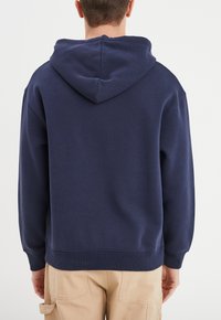 Felpa con cappuccio blu navy realizzata in morbido tessuto, dotata di tasca a marsupio, polsini e orlo a coste, vista da dietro.