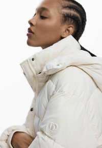 Puffige weiße Jacke mit hohem Kragen, die eine strukturierte Oberfläche, schwarze Knöpfe und ein Logo-Patch am Ärmel aufweist.