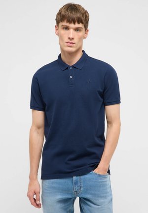 Junger Mann trägt ein dunkelblaues kurzärmeliges Poloshirt und hellblaue Jeans, posiert mit einer Hand in der Hosentasche vor einfachem Hintergrund.