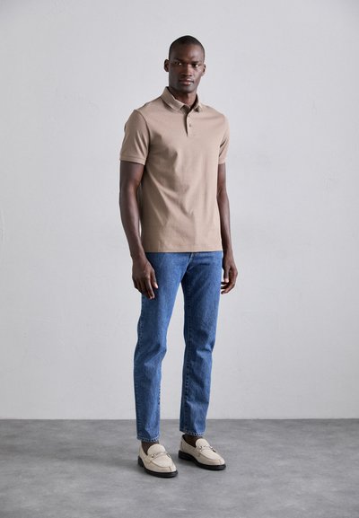 Polo beige à manches courtes, jean droit bleu, et mocassins crème avec semelles noires. Tissu lisse, coupe classique, accents minimaux.