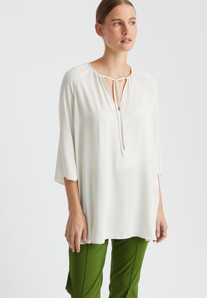 Weiße Bluse mit Rundhalsausschnitt, versehen mit einem Bindedetail und Dreiviertelärmeln, kombiniert mit grünen Hosen. Glatte Textur.