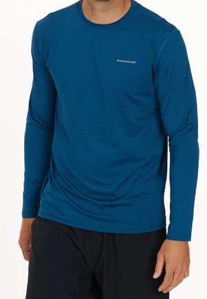 Mann trägt ein eng anliegendes blaues Langarmshirt mit dem „ENDURANCE“-Logo auf der Brust und lockere schwarze Shorts, steht vor einem weißen Hintergrund.
