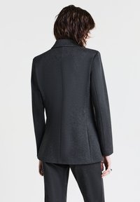 Donna con capelli corti scuri, vestita con un blazer grigio scuro aderente e pantaloni abbinati, in piedi di spalle contro uno sfondo neutro.