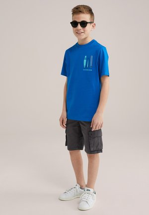 Ljusblå T-shirt med surfbräde grafik, tillsammans med mörkgrå cargo-shorts och vita sneakers. Kort ärm och avslappnad passform.