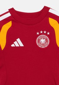 Rdeč športni dres z rumenimi in belimi črtami na ramenih, logotipom Adidas in emblemom Deutscher Fussball-Bund z štirimi zvezdami nad njim.