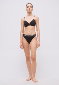 Γυναίκα όρθια, ξυπόλητη, φοράει μαύρο σουτιέν και μαύρο εσώρουχο Polo Ralph Lauren, μπροστά σε απλό λευκό φόντο.