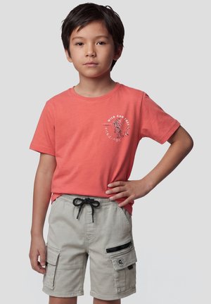 Niño con cabello corto y oscuro, lleva una camiseta coral con un gráfico de tigre y pantalones cortos cargo gris claro, de pie frente a un fondo liso.