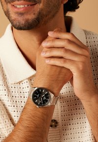 Silver watch with a black dial and silver metal strap. Worn on a tanned wrist, with a textured cream sweater in the background.