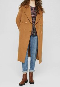 Dubbelzijdige, lange camel tas in zachte stof, met grote knopen, een gekartelde kraag, gecombineerd met een gestreept bovenstuk en blauwe jeans.
