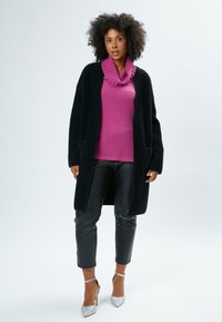 Cardigan lungo nero in peluche con tasche, abbinato a un maglione rosa con collo a cowl, pantaloni in pelle nera e tacchi alti argentati brillanti.