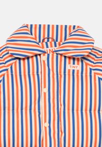 Polstret jakke med stor krage, med vekslende oransje og blå striper på en kremfarget bakgrunn. Inkluderer knappfester og et merke.