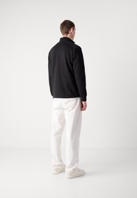 Pulover negru cu guler înalt, văzut din spate, asortat cu pantaloni largi albi și adidași în culori deschise. Textură netedă.