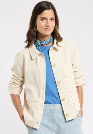 Femme aux cheveux châtains mi-longs portant une veste boutonnée crème, une chemise bleue, une écharpe à motifs et un pantalon rayé, debout les mains dans les poches.