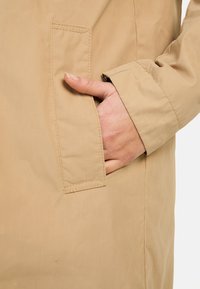 Beige Outdoorjacke mit einer Seitentasche, glatter Textur und genähten Nähten, die ein lässiges Design und eine lockere Passform präsentiert.