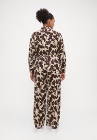 Mono de estampado de vaca marrón y beige con mangas largas y pantalones de corte suelto. Presenta un cinturón en la cintura y un diseño de cuello con solapa.