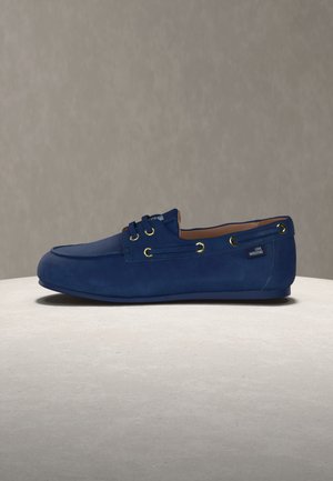 Mocassino in pelle scamosciata blu con occhielli dorati e etichetta "Love Moschino" sul tallone, esposto su una superficie rotonda chiara con sfondo neutro.
