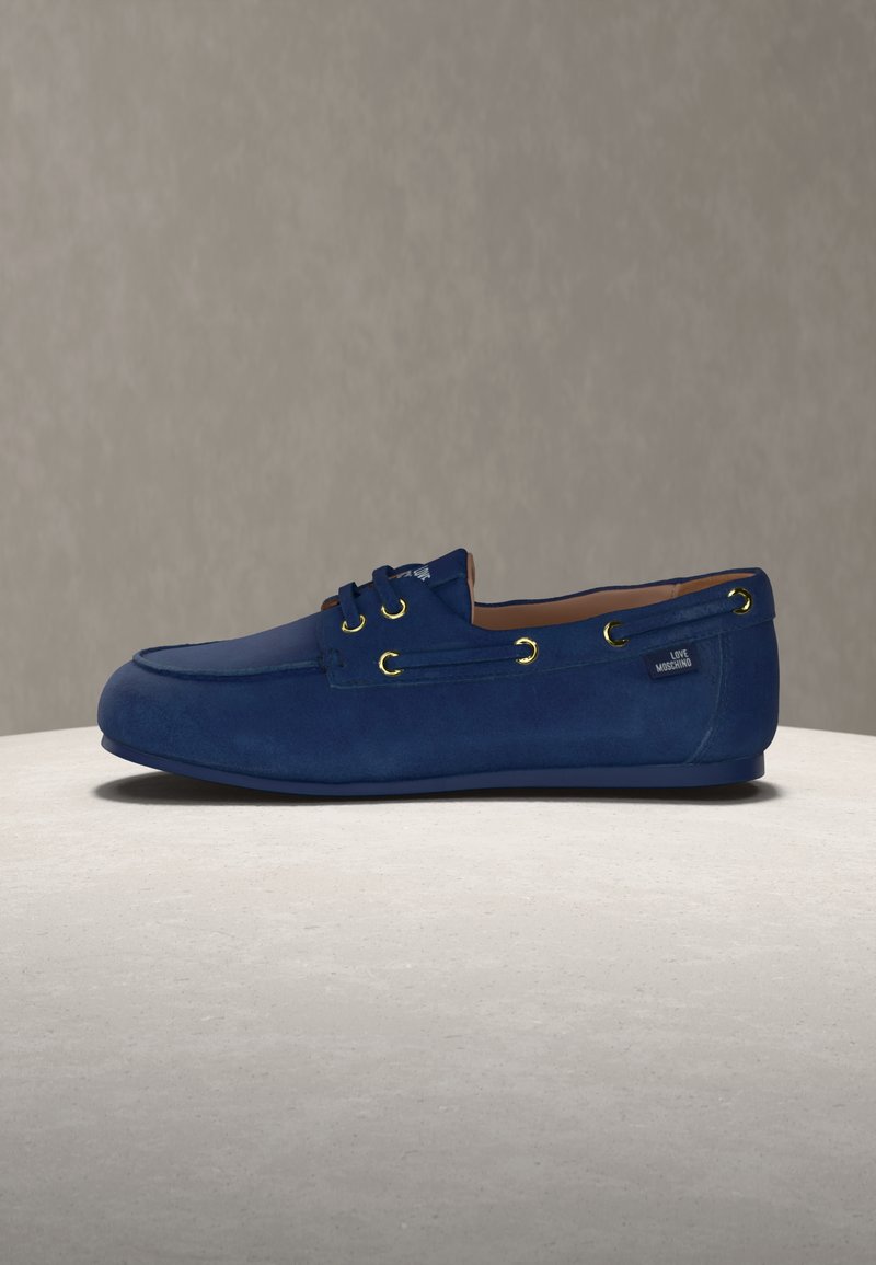 Μπλε μοκασίνι σουέντ με χρυσά τελείωμα και ετικέτα "Love Moschino" στη φτέρνα, τοποθετημένο σε φωτεινή στρογγυλή επιφάνεια με ουδέτερο φόντο.