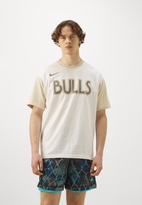 Tricou din bumbac alb cu mâneci bej, având inscripția "BULLS" în litere gri, asociat cu pantaloni scurți cu model black și blue, cu bordură teal.