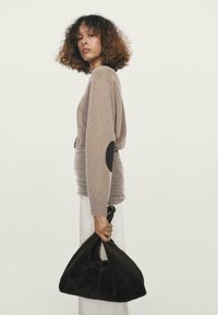 Maglione a maglia color cognac con toppe marroni sui gomiti, abbinato a una borsa in suede marrone scuro, indossato sopra una gonna bianca. La modella è di profilo.