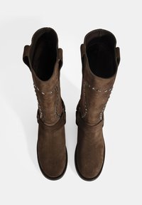 Bottines en daim marron hauteur genou avec des détails décoratifs en œillets argentés et une large ouverture. Semelles plates et accents de boucle sur le côté.