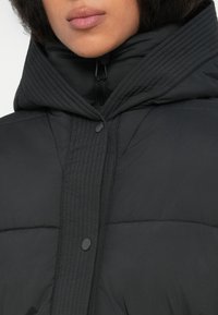Žena nosi crnu zimskojacket s karo uzorkom i visokim ovratnikom, vidljivim patentnim zatvaračem i kopčanjem na dugmadi, usne i donji dio lica djelomično su prikazani.