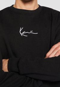 Sudadera negra de tela suave con un logo bordado en blanco en el pecho. Incluye puños y cuello acanalados para un ajuste ceñido.