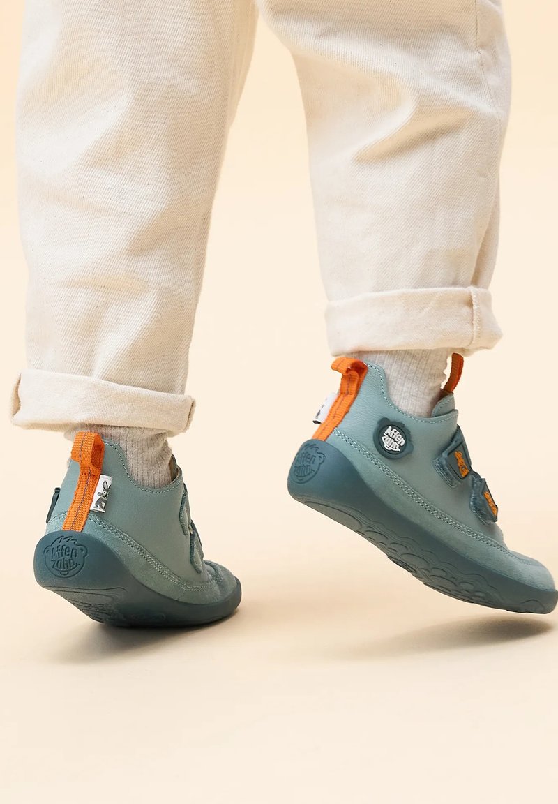 Scarpe flessibili grigio-blu con accenti arancioni, dotate di cinturini con chiusura a strappo, punta arrotondata e suole in gomma testurizzata.