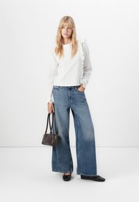 Donna bionda in piedi, con una blusa bianca decorata con pizzo, jeans a gamba larga blu, ballerine nere e in mano una piccola borsa marrone scuro.
