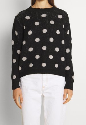 Pull noir avec des pois argentés brillants, col et poignets côtelés, matière tricotée texturée, porté sur un pantalon blanc.