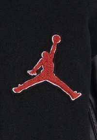 Tissu noir avec un logo Jumpman brodé texturé en rouge. Le logo présente une silhouette dans une pose de saut, entourée de blanc.