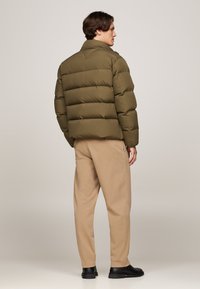 Doudoune vert olive avec col montant, design matelassé et coupe décontractée, associée à un pantalon beige et des chaussures noires.