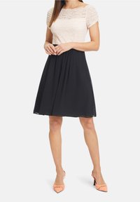 Kobieta w czarnej spódnicy do kolan, z białą koronkową bluzką z krótkim rękawem i koralowymi, plecionymi czółenkami z szpiczastym noskiem, stoi z rękami złożonymi.