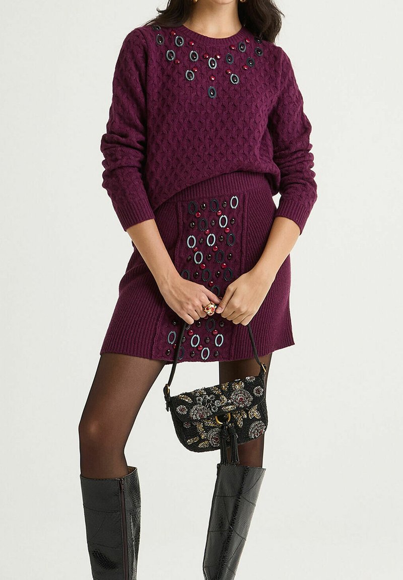 Pull violet en maille avec un motif texturé et des accents de bijoux. Jupe assortie violette avec des ornements remarquables. Sac à main noir orné.