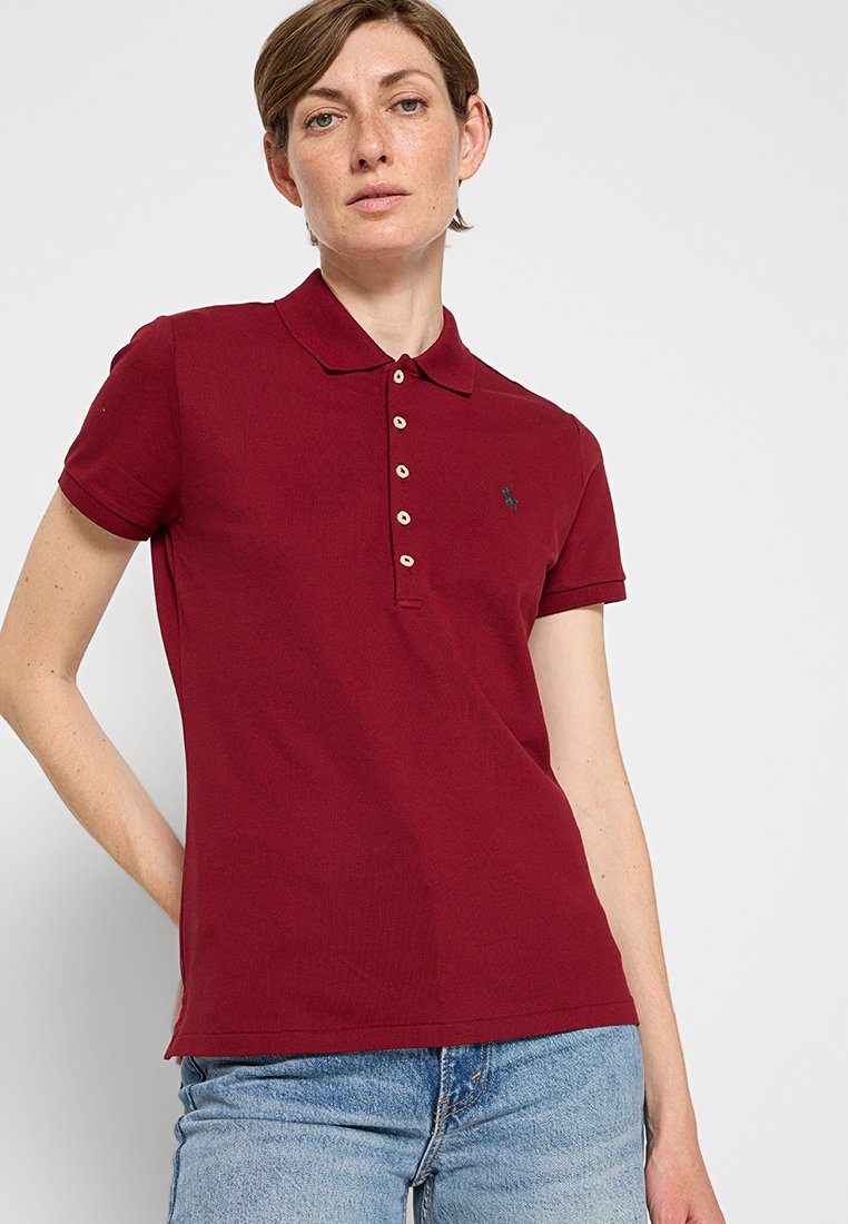 Burgundowa koszulka polo z kołnierzykiem, krótkimi rękawami i listwą z małymi guzikami, w połączeniu z niebieskimi dżinsami. Logo na lewej piersi.