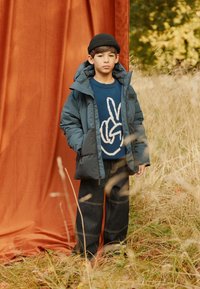 Puffy teal jacket with a hood, layered over a blue sweater featuring a white hand gesture. Dark denim pants with visible stitching details.