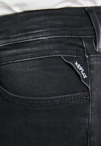 Black denim jeans featuring a rear pocket with a blue Replay label, contrasting white stitching, and a smooth texture.