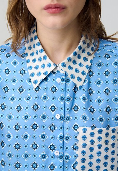 Camicia a righe blu con una base azzurra chiara e motivi floreali blu scuro. Colletto bianco con una stampa floreale più piccola. Chiusura con bottoni davanti.