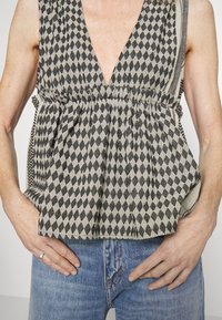 Top de patrón negro y crema con un escote en V profundo, con formas de diamante y cintura plisada, combinado con jeans de mezclilla azules.