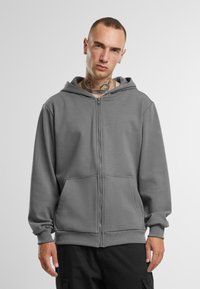 Grå zip-hoodie med kængurulomme, ribbede manchetter og en rummelig hætte, lavet af blødt stof med en glat tekstur.