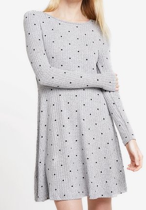 Femme portant une robe grise en maille côtelée à manches longues avec des pois noirs, arrivant mi-cuisse.